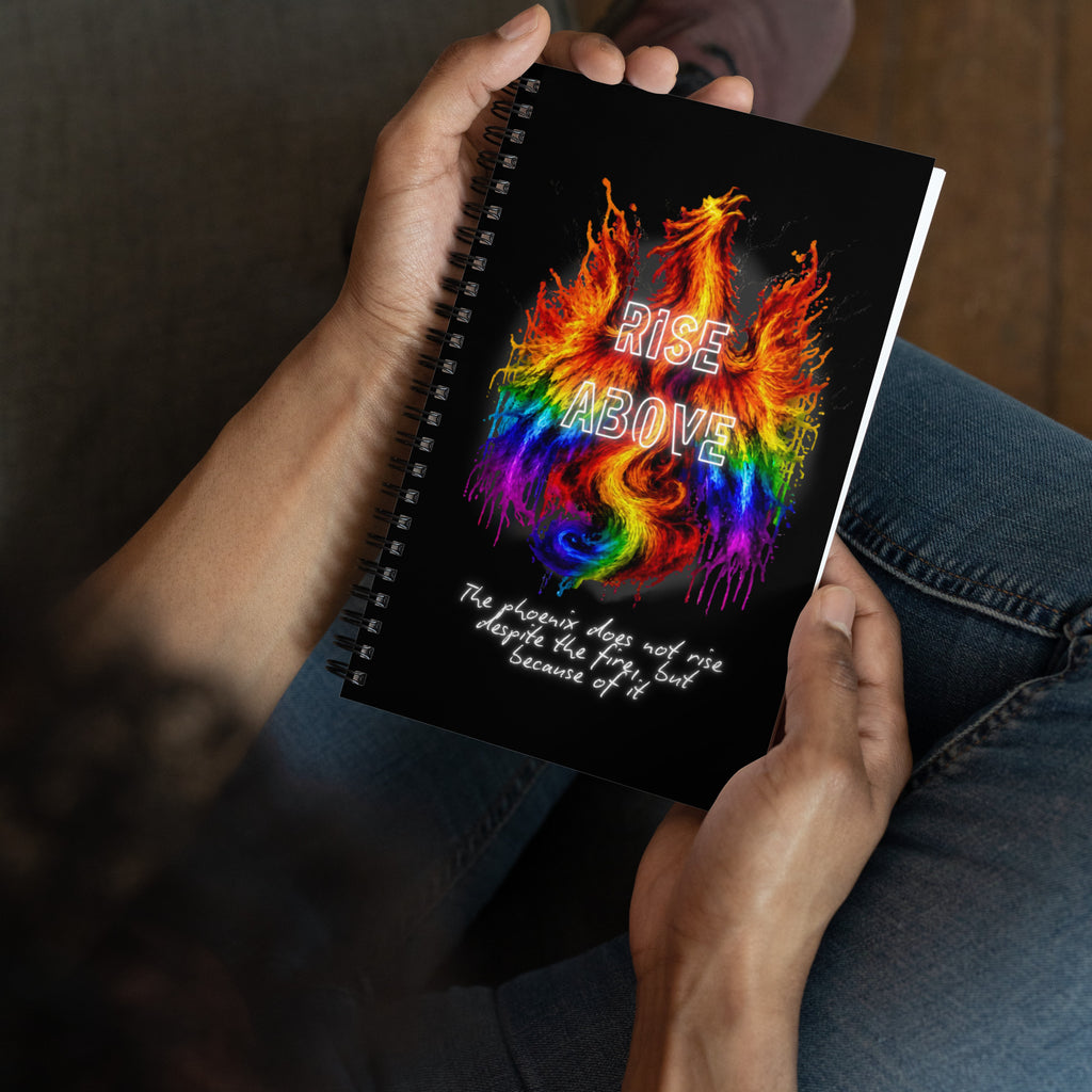 A person holding a spiral-bound notebook with a black cover featuring a colorful rainbow dragon design and the words 'RISE ABOVE' in white. The person is wearing blue jeans and sitting indoors.