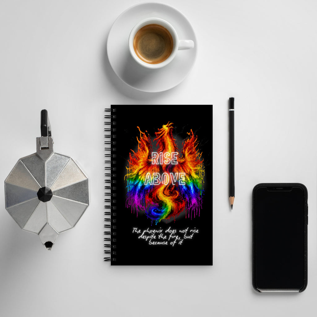 A spiral-bound notebook with a black cover and a colorful rainbow phoenix graphic reading 'RISE ABOVE' is arranged in a flat lay on a light gray surface, surrounded by a cup of coffee, a moka pot, a pencil, and a smartphone.